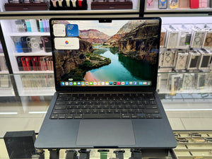A dark gray Apple MacBook laptop with its screen turned on, displaying a canyon landscape wallpaper and dock icons, set on a glass display counter with store shelves in the background.