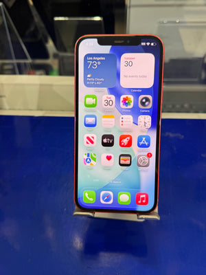 A red iPhone with its screen turned on, displaying the home screen with various app icons, resting on a small metal stand on a blue surface.
