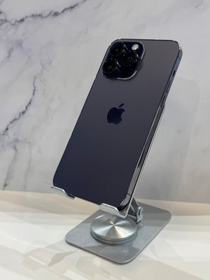 A dark blue-gray Apple iPhone 12 Pro displayed upright on a silver metal stand, showing the back of the phone with the Apple logo and triple camera array against a white marble-patterned background.