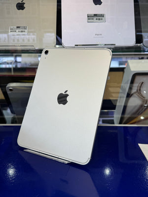 A silver Apple iPad Pro shown from the back, standing on a display stand on a dark blue reflective surface, with Apple products visible in the background.