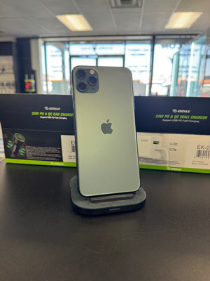An Apple iPhone with a light green back is shown from the rear, resting on a dark gray stand on a dark countertop. Its retail packaging is visible in the background.