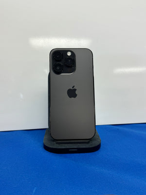 A black Apple iPhone standing upright on a small black charging stand, photographed against a white wall with a blue surface below.