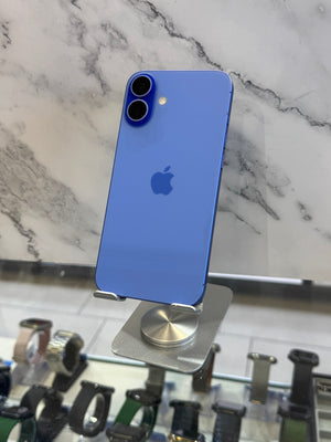 A blue Apple iPhone shown from the back, displayed on a metal stand in front of a marble-patterned wall. Other watches are visible on a counter below.