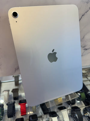 An Apple iPad with a light silver aluminum back, featuring the iconic Apple logo in the center and a single rear camera lens. The device is photographed at an angle in what appears to be a retail or showroom setting, with a marble-patterned wall in the background and a glass display case containing various smartwatches below.