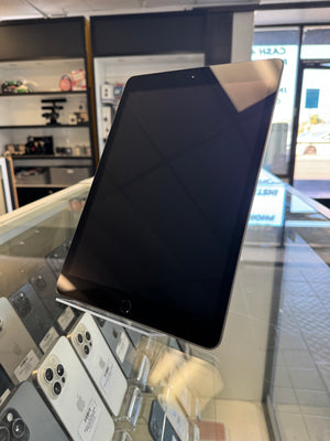 A black tablet device with a glossy screen, standing on a clear glass display counter inside a retail store. Other phones are visible in the background on the counter.