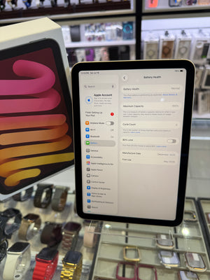 An iPad displaying the battery health settings screen, photographed in a retail store setting with watch displays and an Apple product box visible in the background.