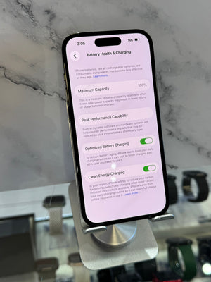 A black smartphone displayed on a stand, showing the battery health and charging settings screen on its screen. The phone is photographed in a real environment with a marble-like surface and other devices blurred in the background.