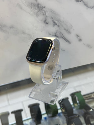 A smartwatch with a cream-colored sport band and a gold-toned case, displayed on a clear acrylic stand in front of a white marble-patterned background.