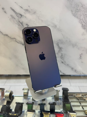 A dark blue-to-purple gradient iPhone 12 Pro Max displayed on a silver stand, with the Apple logo visible on the back. The phone is photographed in a retail or showroom setting with a marble-patterned wall and watch displays in the background.