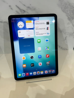 An iPad with a black bezel displaying the iOS home screen, resting on a clear acrylic stand on a light beige surface against a white marble-patterned background.
