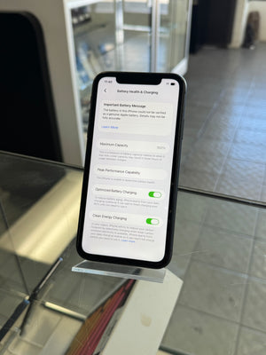 A black smartphone displayed upright on a clear acrylic stand on a glass table, showing the Settings screen for Battery Health & Charging in a real indoor environment with blurred background elements.